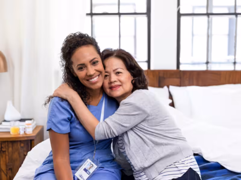 A nurse in blue scrubs sits on a bed, smiling as an older woman hugs her. They both look happy in a bright, cozy room with large windows in the background.