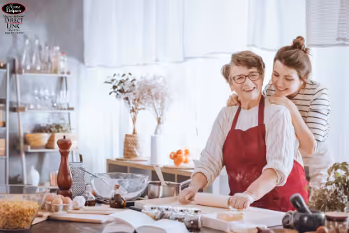 Two women in a bright kitchen smile as one, wearing an apron, rolls dough while the other hugs her from behind. Baking ingredients and utensils are spread on the counter.