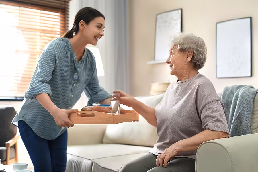 A young woman smiles as she brings a tray with food and a glass of water to an older woman sitting on a couch in a cozy living room. The older woman looks up at her with a grateful expression.