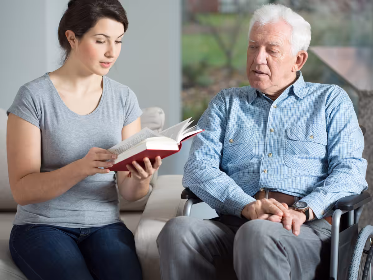 A young woman reads a book to an elderly man in a wheelchair.