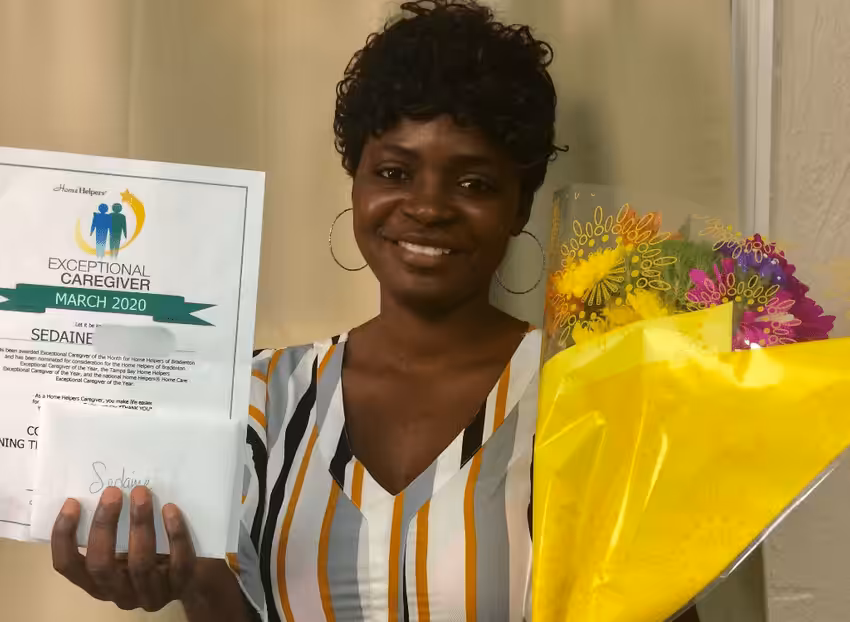 A smiling woman holds a bouquet of flowers and a certificate for Exceptional Caregiver March 2020. She wears a striped shirt and stands in front of a beige background.