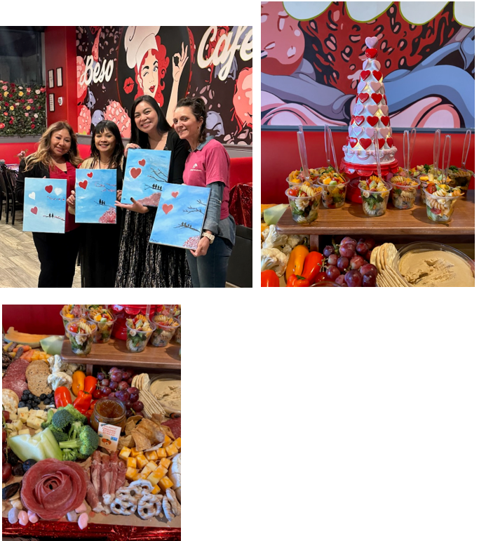 Four women smile holding painted canvases with heart designs in front of a colorful mural; nearby, there are displays of fresh fruit cups, snacks, and a charcuterie board with cheeses, meats, grapes, and crackers.