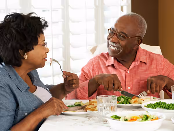 An older couple sits at a dining table, smiling and enjoying a meal together. The man cuts his food while the woman holds a fork. Plates of vegetables and glasses of water are on the table in a bright, sunlit room.