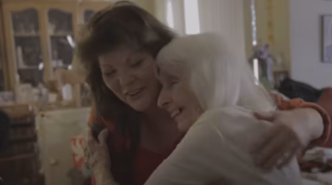 Two women, one younger and one older with white hair, are smiling and embracing each other warmly in a cozy home setting.
