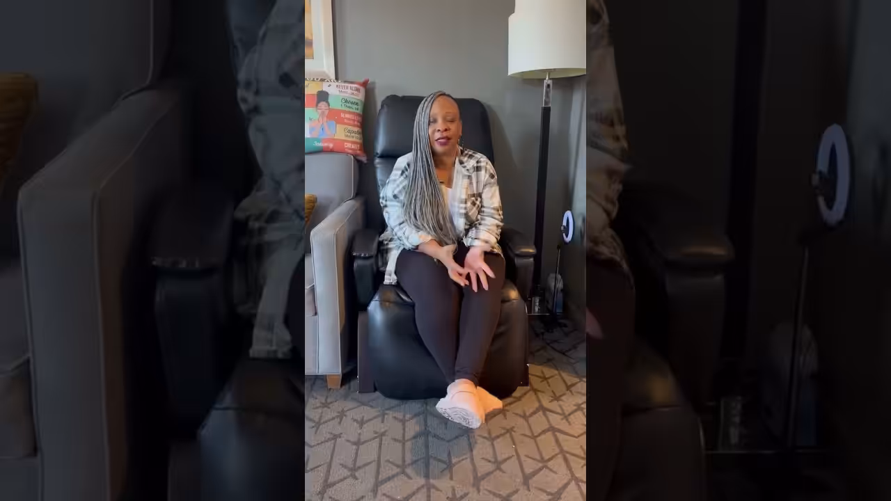 A person with long braided hair, wearing a plaid shirt, black leggings, and light slippers, sits on a black chair with hands resting on their lap in a cozy, carpeted room. A floor lamp and a colorful pillow are nearby.