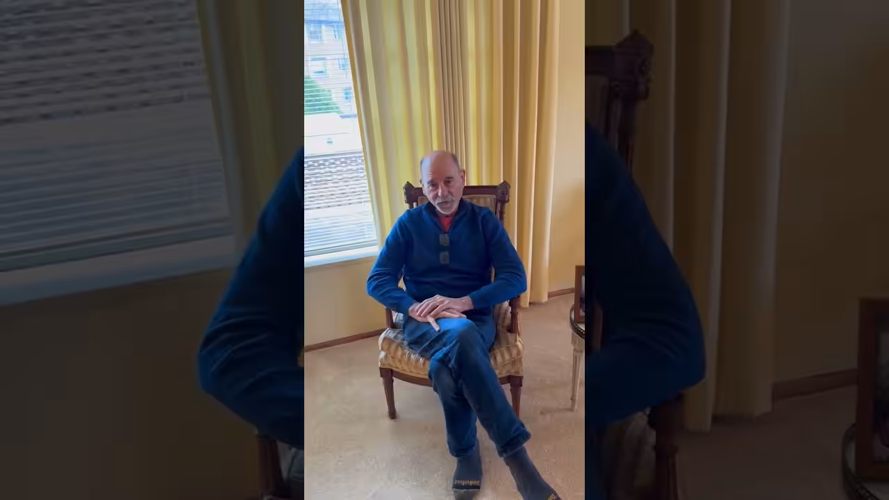 A man with a bald head and gray beard, wearing a blue sweater and jeans, sits on a wooden chair in a room with yellow walls and curtains, next to a window with blinds.