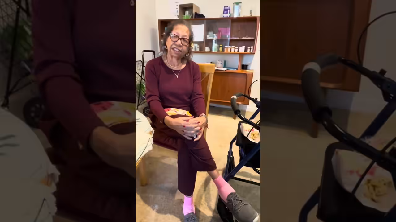 An elderly woman with glasses, wearing a maroon outfit and pink socks, sits on a chair indoors. A walker is nearby, and there is a cabinet with bottles and books in the background.