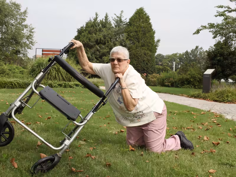 An elderly woman wearing sunglasses is kneeling on the grass in a park while holding onto a walker that has tipped over. She appears to be trying to get up or call for help. Trees and greenery are in the background.