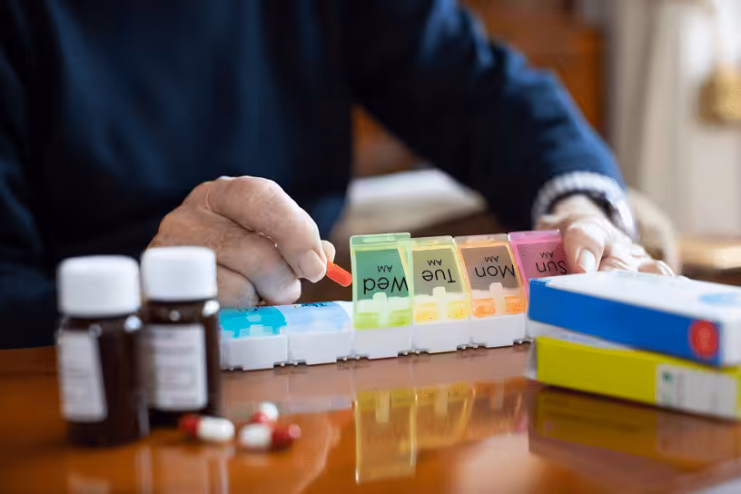 An older adult’s hands organizing pills into a weekly pill organizer, with medicine bottles, blister packs, and loose capsules on a wooden table.