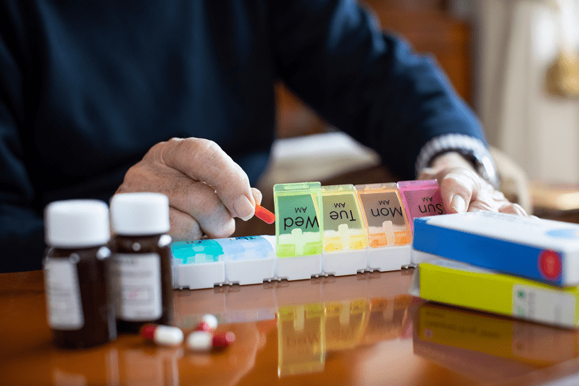 An older adult’s hands organizing pills into a weekly pill organizer, with medicine bottles, blister packs, and loose capsules on a wooden table.