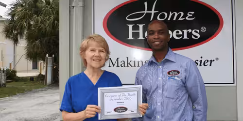Two people stand smiling outside a Home Helpers building. The woman on the left holds a certificate for Caregiver of the Month September 2016. The man on the right stands beside her, both wearing work uniforms.