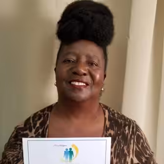 A woman with a high updo hairstyle and patterned brown top smiles while holding a certificate in front of a neutral curtain background.