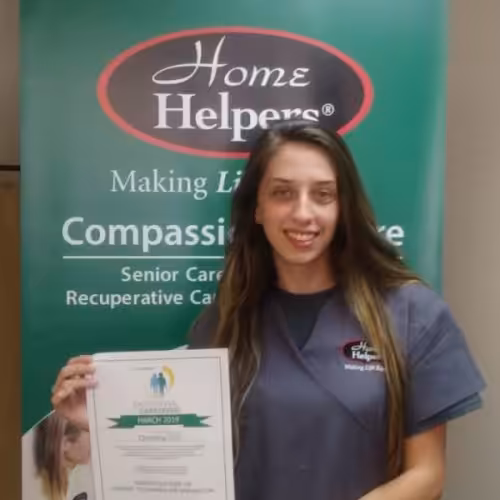 A smiling woman in gray scrubs holds a certificate, standing in front of a green Home Helpers banner promoting compassionate senior care.
