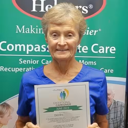 An older woman with short hair, wearing blue scrubs, smiles while holding an Exceptional Caregiver June 2019 certificate in front of a banner that says Compassionate Care.