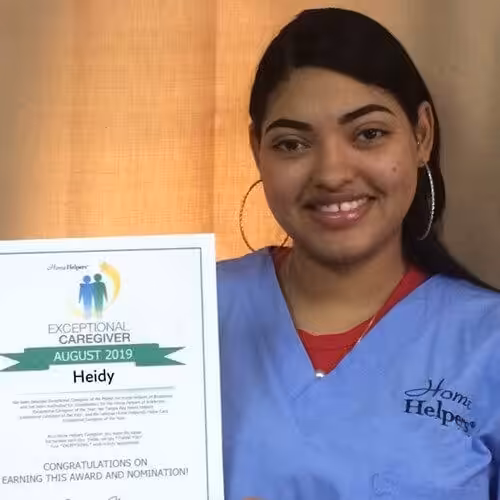 A woman wearing blue scrubs and hoop earrings smiles while holding an Exceptional Caregiver August 2019 award certificate with the name Heidy printed on it. A beige curtain is in the background.