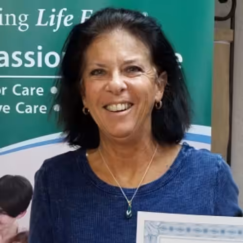 A woman with dark hair wearing a blue top and a necklace smiles at the camera, standing in front of a green and white banner with text. She is holding a certificate.