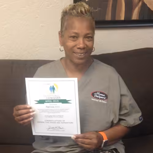 A woman wearing gray scrubs with a Home Helpers logo sits on a couch and smiles, holding a certificate for Exceptional Caregiver April 2019.
