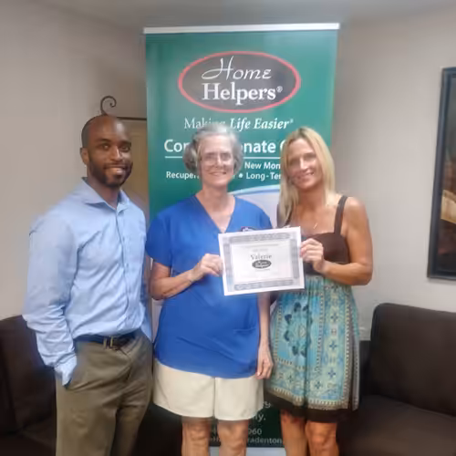 Three people stand indoors in front of a Home Helpers banner. The woman in the center, holding a certificate, is flanked by a man on the left and a woman on the right. All three are smiling.
