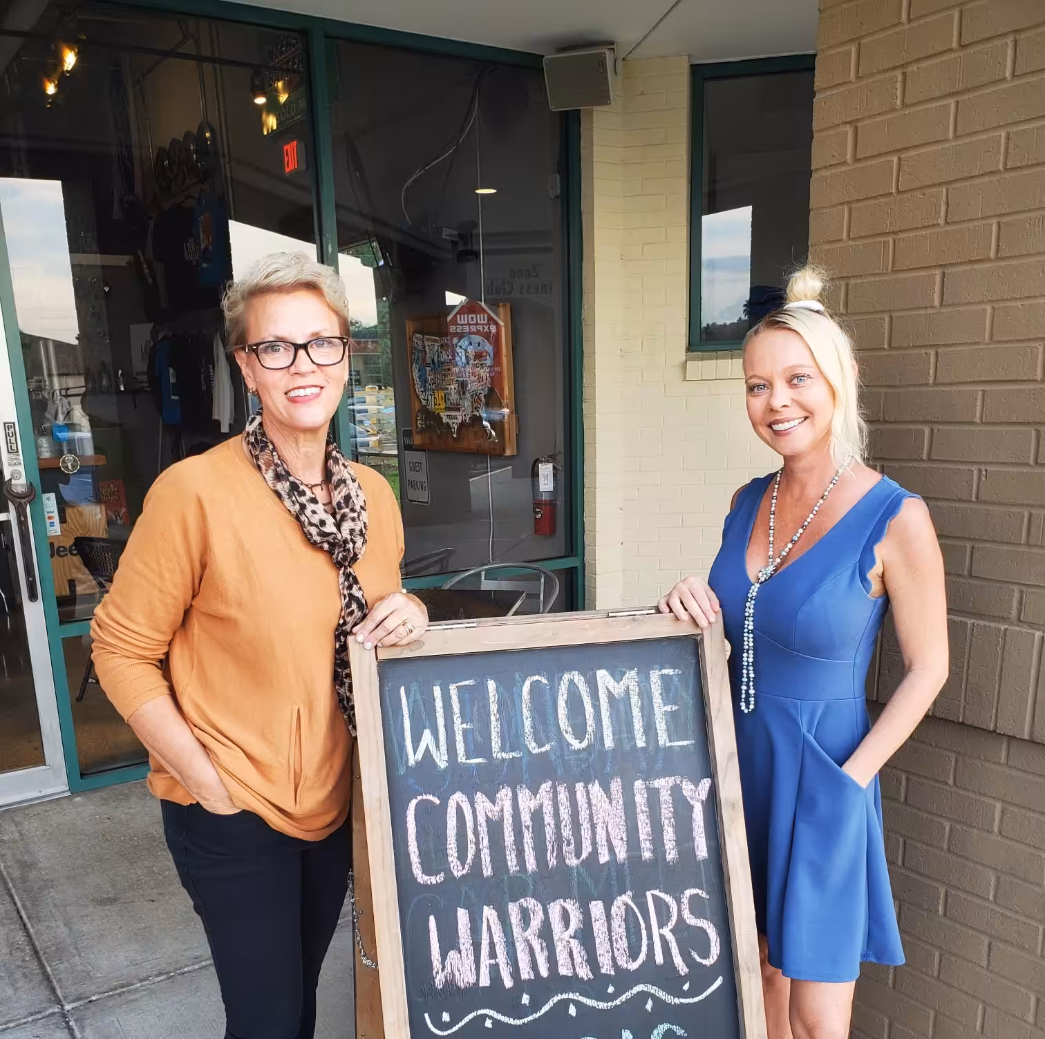 Two women stand outside a storefront next to a chalkboard sign that reads “Welcome Community Warriors.” One woman wears an orange sweater and scarf; the other wears a blue dress and necklace.
