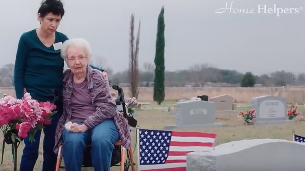 An elderly woman in a wheelchair, accompanied by a younger woman, sits beside a grave adorned with flowers and an American flag in a cemetery. The Home Helpers® logo appears in the top right corner.