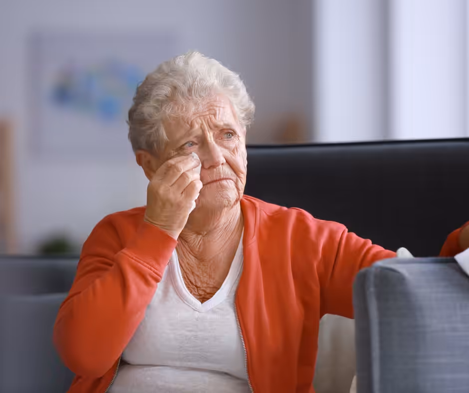 An elderly woman with short gray hair, wearing a white shirt and orange cardigan, sits on a couch wiping tears from her face, appearing sad or emotional. The background is softly blurred.