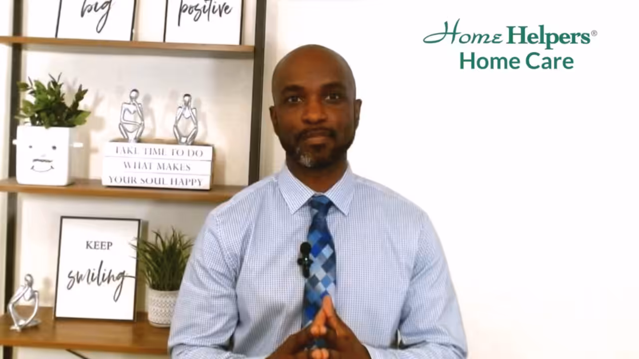 A man in a blue shirt and tie sits in front of shelves with décor and motivational quotes. The Home Helpers Home Care logo appears in the top right corner.