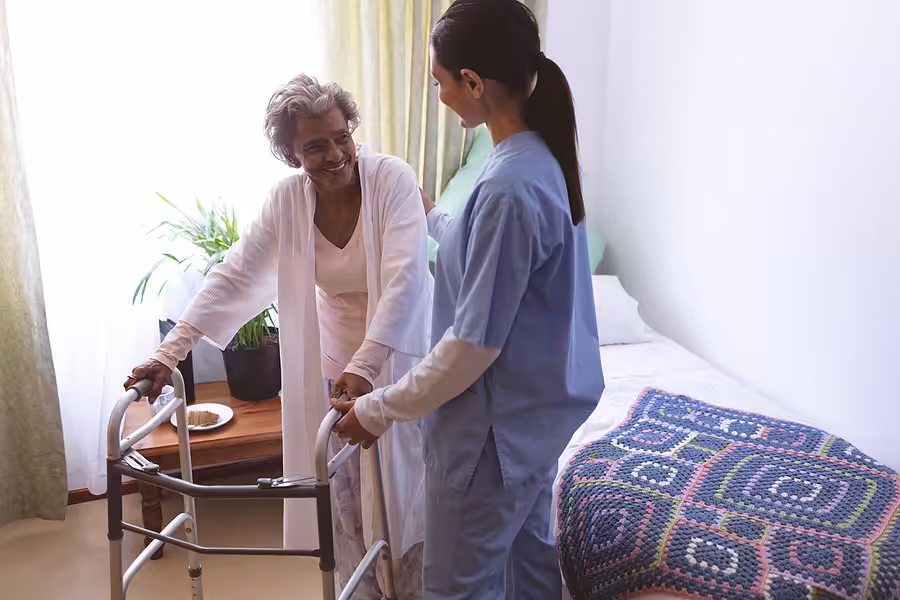 An elderly woman using a walker smiles at a nurse standing beside her in a bright bedroom. The bed has a colorful crocheted blanket, and sunlight streams through a window with light curtains.