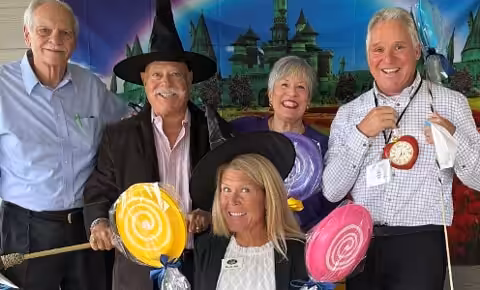 Five adults dressed in Wizard of Oz themed costumes, holding large colorful lollipops and props, pose and smile in front of a backdrop depicting the Emerald City.