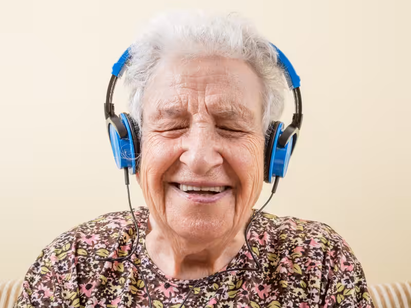 An elderly woman with short white hair smiles with her eyes closed while wearing blue headphones and a floral-patterned top, sitting against a light background.