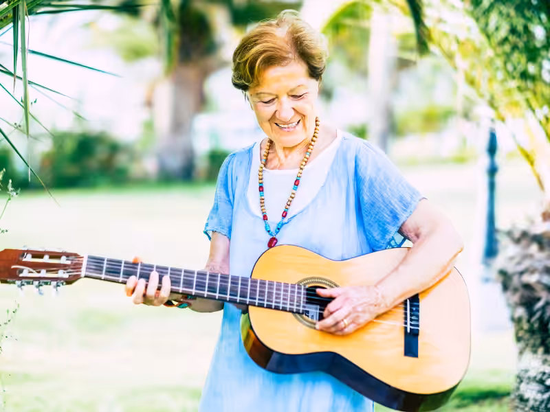 An older woman in a light blue dress plays an acoustic guitar outdoors, smiling and standing among green plants and trees on a sunny day.