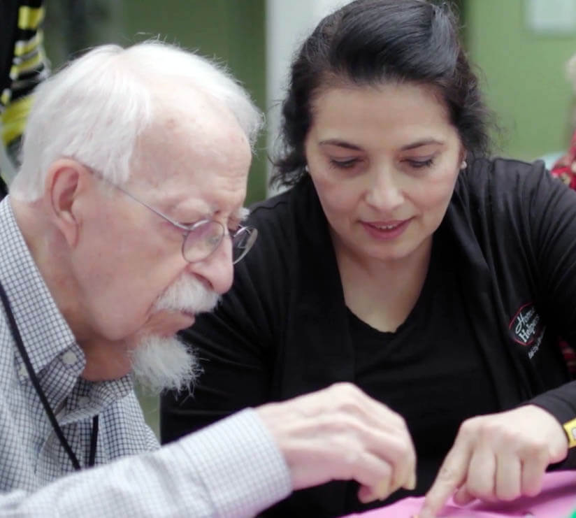 An elderly man with white hair and glasses sits with a woman who points at something in front of them; they appear to be concentrating on an activity together.
