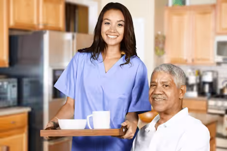A smiling caregiver in scrubs holds a tray with a cup and bowl, standing next to an older man who is sitting and smiling in a kitchen setting.