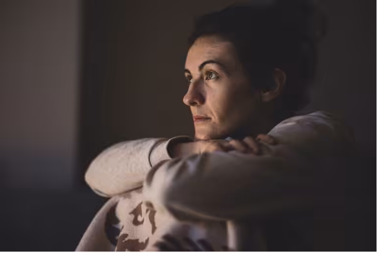A woman sits indoors with her arms resting on her knees, gazing thoughtfully into the distance. Soft lighting highlights her contemplative expression against a dark, blurred background.