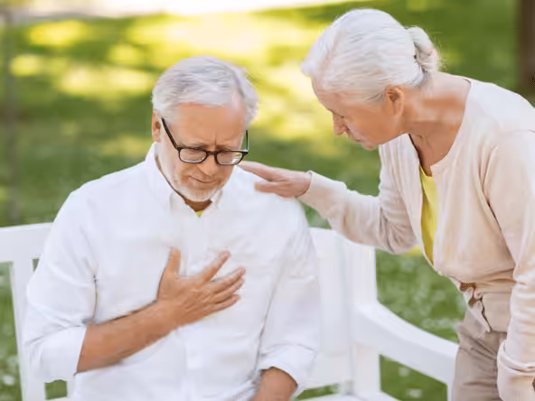 An elderly man sits on a bench holding his chest in discomfort while an elderly woman stands beside him, placing a comforting hand on his shoulder. They are outdoors in a park-like setting.