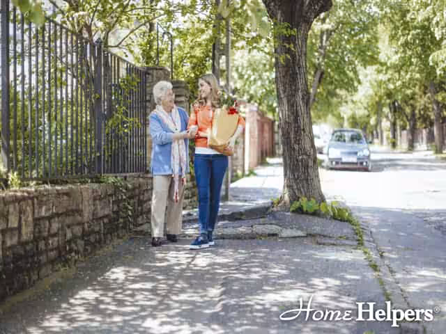 A young woman helps an elderly woman walk along a shaded sidewalk, carrying a grocery bag. Trees and a car line the street. Home Helpers logo appears in the bottom right corner.