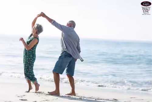 An older couple dances joyfully on a sandy beach near the shoreline, with the ocean waves gently rolling in the background and sunlight illuminating the scene.