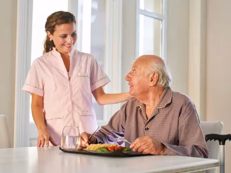 A smiling caregiver stands beside an elderly man sitting at a table with a meal tray and a glass of water, both looking at each other warmly in a bright, homey setting.