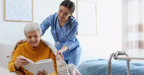 A nurse in blue scrubs smiles while placing a blanket over an elderly woman sitting and reading a book in a cozy, well-lit room. A walker and a bed are visible in the background.