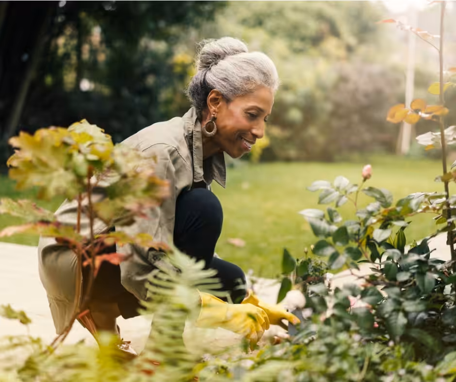 An older woman with gray hair, wearing gardening gloves and a khaki jacket, kneels outdoors while tending to plants and flowers in a lush, green garden.