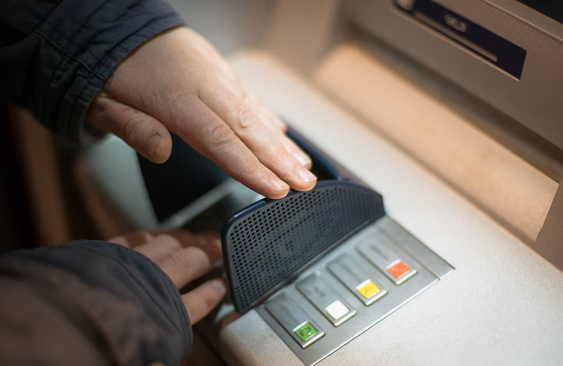 A person shields the keypad with one hand while entering a PIN at an ATM, protecting their code from being seen. Close-up of hands and ATM buttons.
