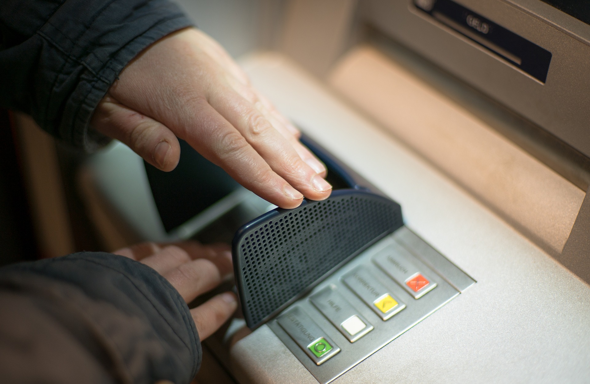 A person shields the keypad with one hand while entering a PIN at an ATM, protecting their code from being seen. Close-up of hands and ATM buttons.