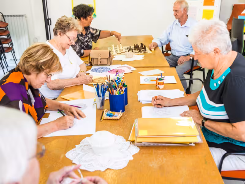 A group of elderly people sit around a table engaged in various activities such as drawing, coloring, and playing chess in a bright, communal room. Art supplies and papers are spread across the table.
