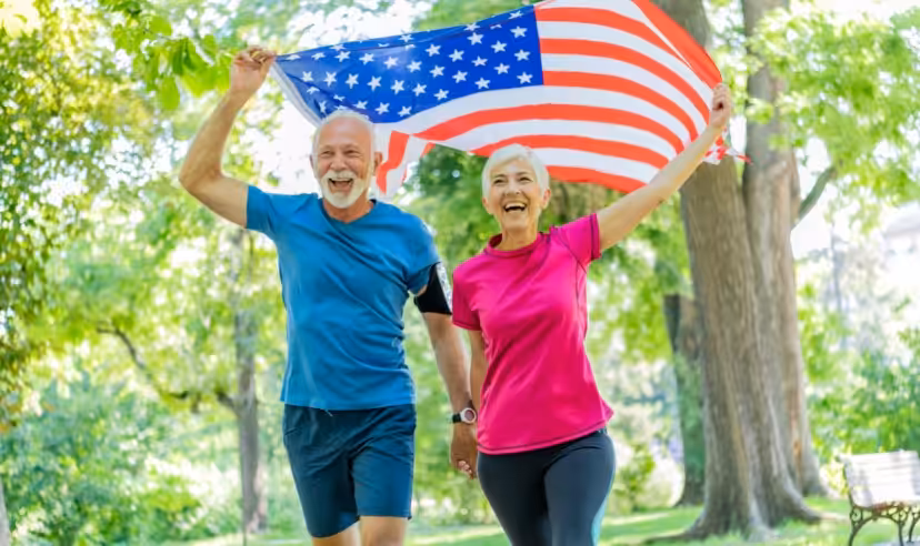 An older couple in athletic clothes smiles and walks outdoors while holding up an American flag, surrounded by green trees and sunlight in a park.