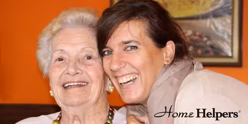 An elderly woman and a younger woman smiling together indoors with an orange wall and framed picture behind them. The Home Helpers logo is in the bottom right corner.