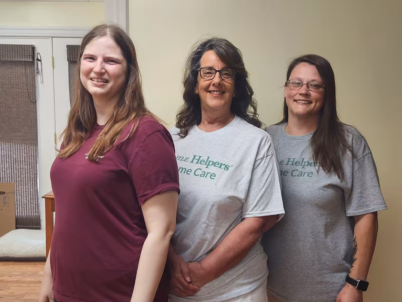 Three women stand indoors, smiling at the camera. Two wear matching gray Heavenz Helpers Home Care t-shirts, while the third wears a maroon t-shirt. They are standing in front of a pale yellow wall and a window.