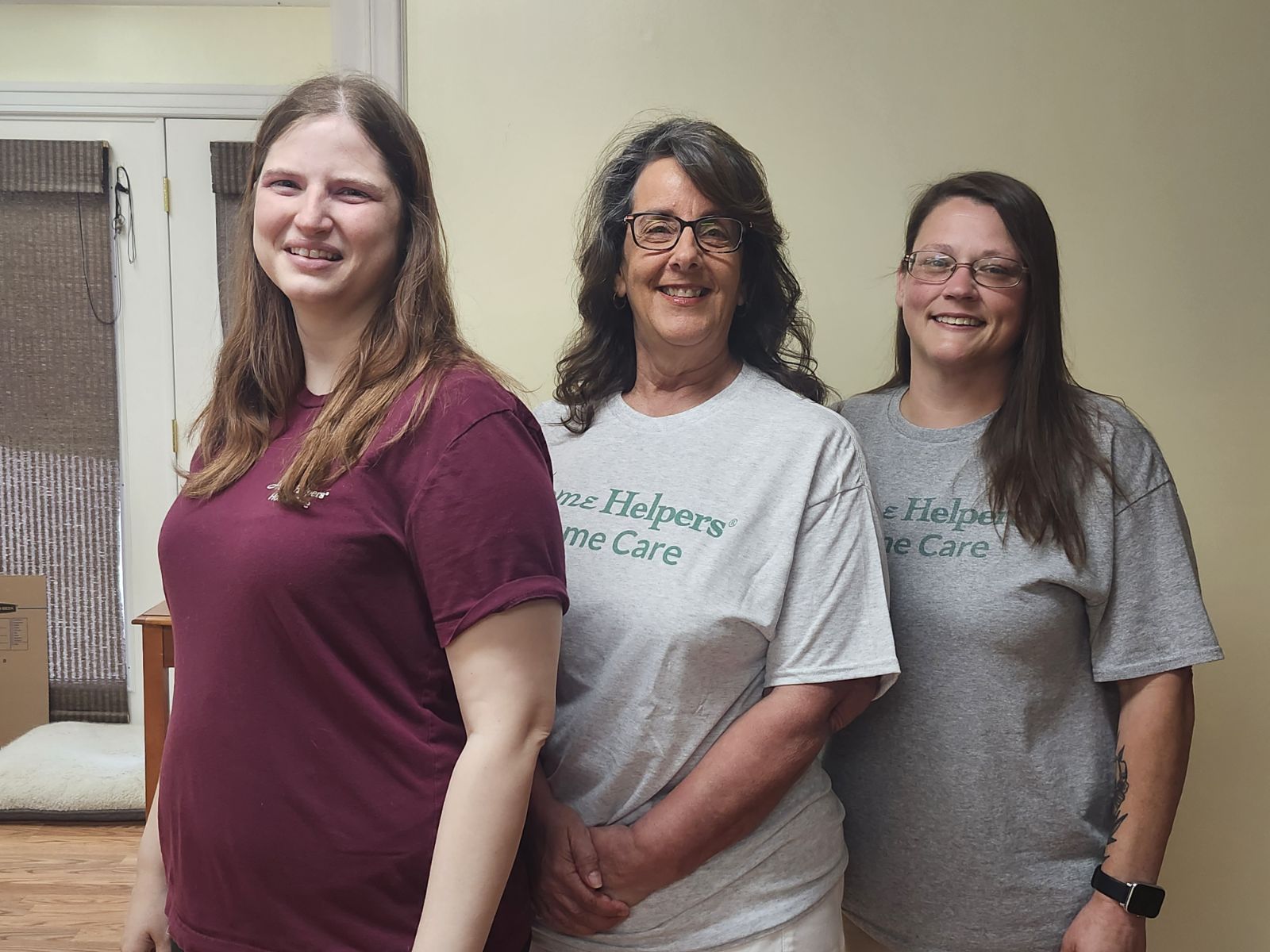 Three women stand indoors, smiling at the camera. Two wear matching gray Heavenz Helpers Home Care t-shirts, while the third wears a maroon t-shirt. They are standing in front of a pale yellow wall and a window.