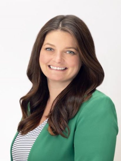 A woman with long brown hair, wearing a green blazer over a white and black striped shirt, smiles at the camera against a plain white background.