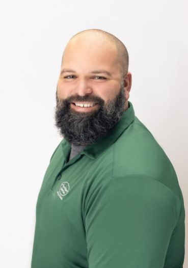 A bearded man with a shaved head, wearing a green collared shirt, smiles while posing against a plain white background.