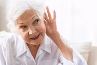 An elderly woman with gray hair in a white shirt is smiling and holding her hand to her ear, as if trying to hear better. She is seated in a well-lit room with a soft background.
