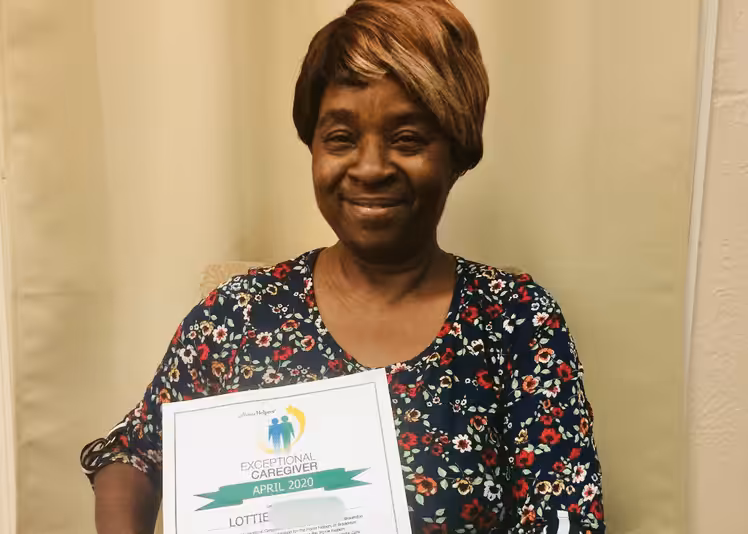 A smiling woman with short brown hair, wearing a floral dress, holds a certificate titled Exceptional Caregiver April 2020 while sitting in front of a beige curtain.